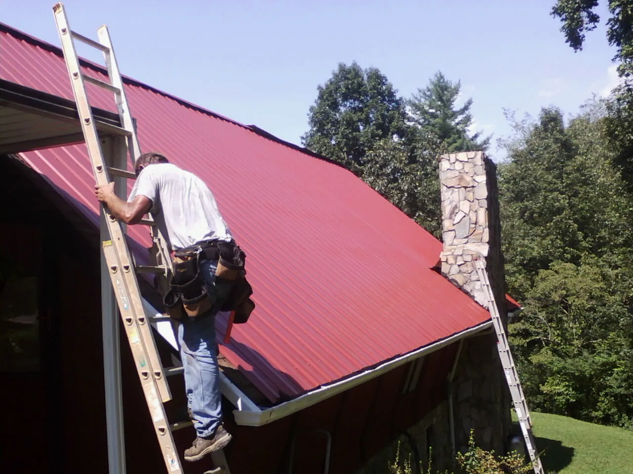 Roofer on a ladder beside a red metal roof and stone chimney — Soffit & Fascia Repair by Roofing Brothers in New Marshfield